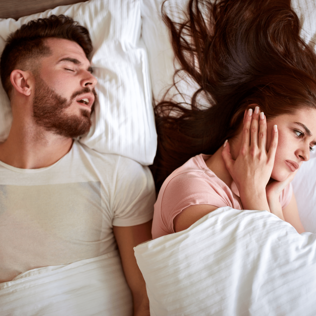A man is lying in bed on his back, snoring loudly with his mouth open. Beside him, his wife is sitting up in bed, looking frustrated and tired, with her hand over her ear and a frown on her face.